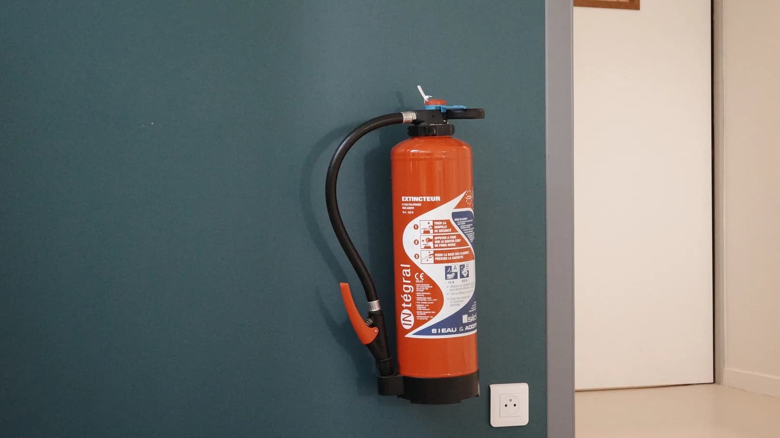 Extincteur à poudre rouge fixé au mur d'un établissement recevant du public, à côté d'une porte